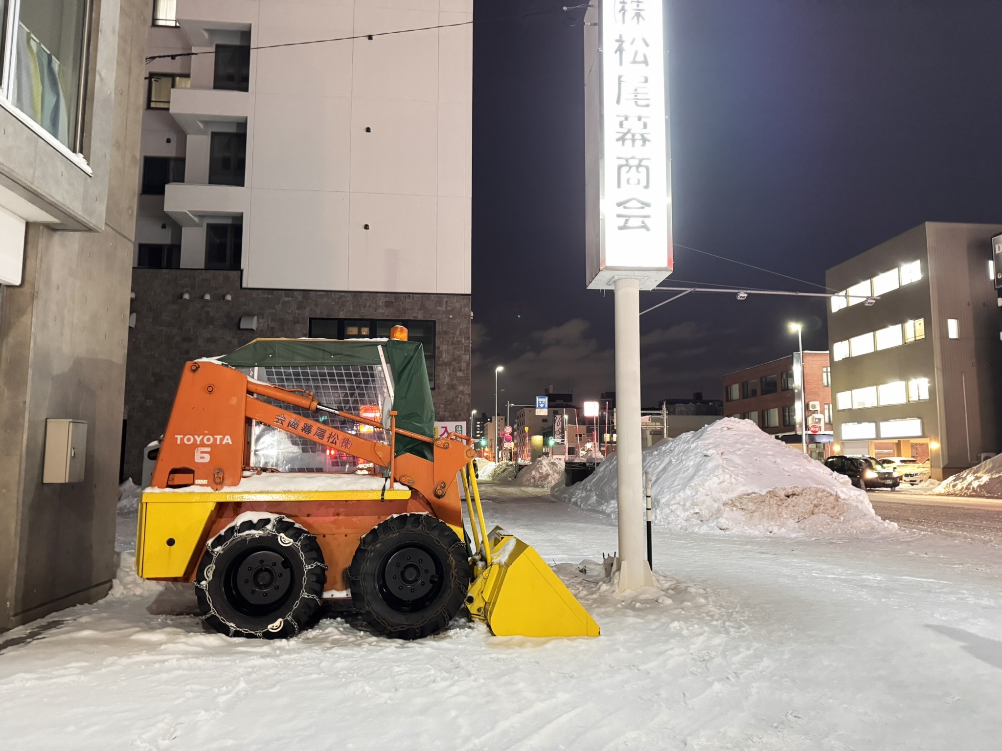 Snow-covered Japanese street scene with a loader and illuminated sign.